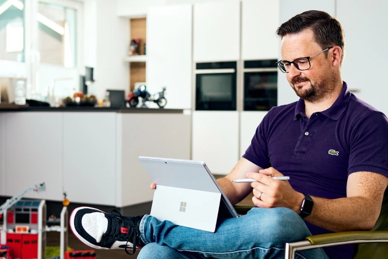 Mann arbeitet entspannt mit Tablet und Stylus im modernen Homeoffice mit Küche im Hintergrund.